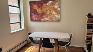 Ein Zimmer mit weißem Tisch, zwei Stühlen, einem großen abstrakten Gemälde, einem Fenster und einem Bücherregal.