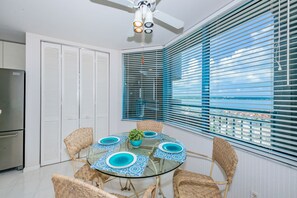 A dining area with a glass table, wicker chairs, and a view through blue blinds. - WHITE SANDS BEACHFRONT SUNSET SUNRISE LUX EVERY ROOM SEA POOLS SPAS IN/OUT (Clearwater)