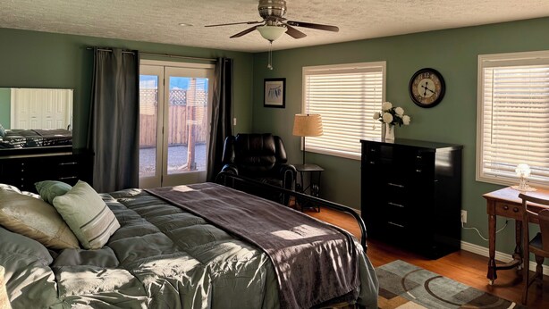 A bedroom with a bed, a desk, a chair, a dresser, a ceiling fan, a clock, and a window with blinds.