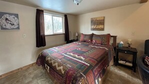 A bedroom with a large bed, a window with curtains, a ceiling fan, a bedside table with a lamp, and a wall-mounted artwork. - Peaceful 1-bedroom cabin in charming Talkeetna perfect for getaway (Talkeetna)