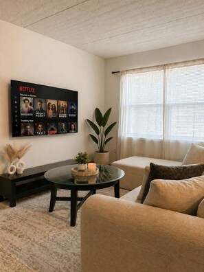 Ein Wohnzimmer mit Flachbildfernseher, der Inhalte von Netflix zeigt, einem runden Glastisch, einer Couch, einer Topfpflanze und einem Fenster mit durchsichtigen Vorhängen.