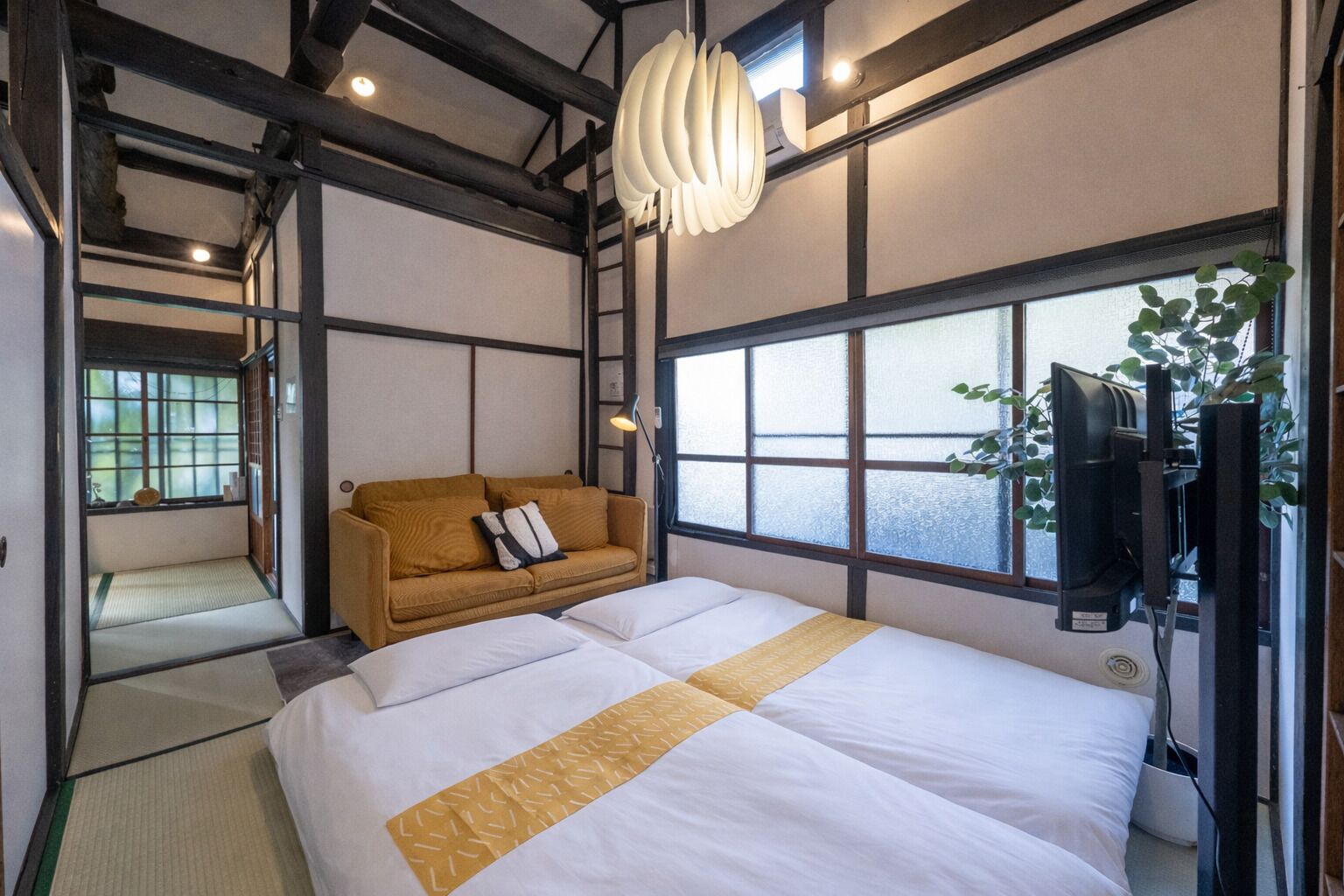 A traditional Japanese-style room with tatami flooring, a bed with white bedding and a yellow accent, a brown sofa with cushions, a television, and a large window with a view.