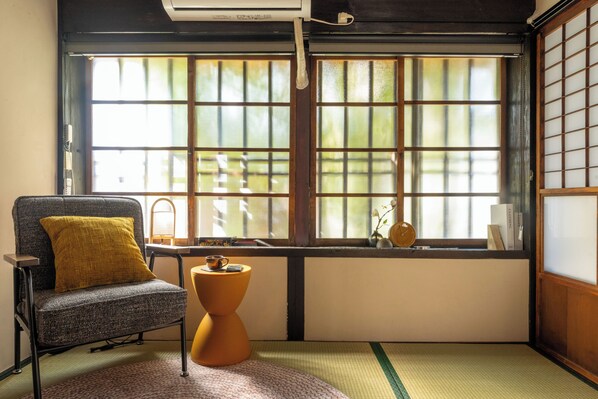 A room with tatami flooring, a traditional Japanese-style chair, a small table with a cup, and a window with a view of greenery.