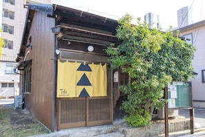 A traditional Japanese building with a wooden exterior, a yellow entrance sign, and a small garden area.