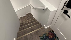 Una scala con gradini in legno che conduce a una piattaforma con uno scaffale bianco, due piante in vaso e una presa elettrica sulla parete.