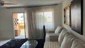 A living room with a sofa, a glass coffee table, a ceiling fan, a window with curtains, and a framed picture on the wall. - Huntington Beach Condo (Huntington Beach)