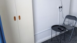 A room with a white wardrobe, a small round table with a remote, a grey chair, and a TV mounted on the wall. - Studio Sleeps 2 with Parking (Oldbury)