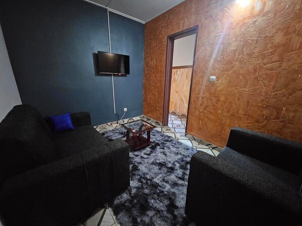 A room with a flat-screen TV mounted on the wall, a dark-colored sofa with a blue pillow, and a patterned rug on the floor.