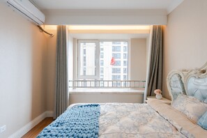 A bedroom with a bed, a window with curtains, a balcony, and a view of buildings.