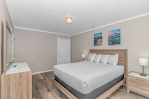 Un dormitorio con cama, mesita de noche, cómoda, puerta y dos cuadros colgados en la pared.