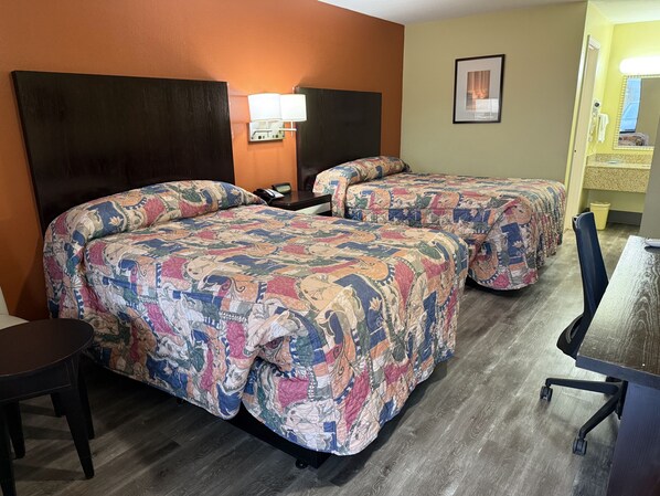 A hotel room with two beds, a desk, a chair, a lamp, and a framed picture on the wall.
