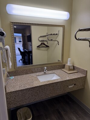 Un bagno d'hotel con un piano di lavoro in granito, un lavandino, uno specchio, un asciugacapelli, un portasciugamani e un asciugamani ad aria.