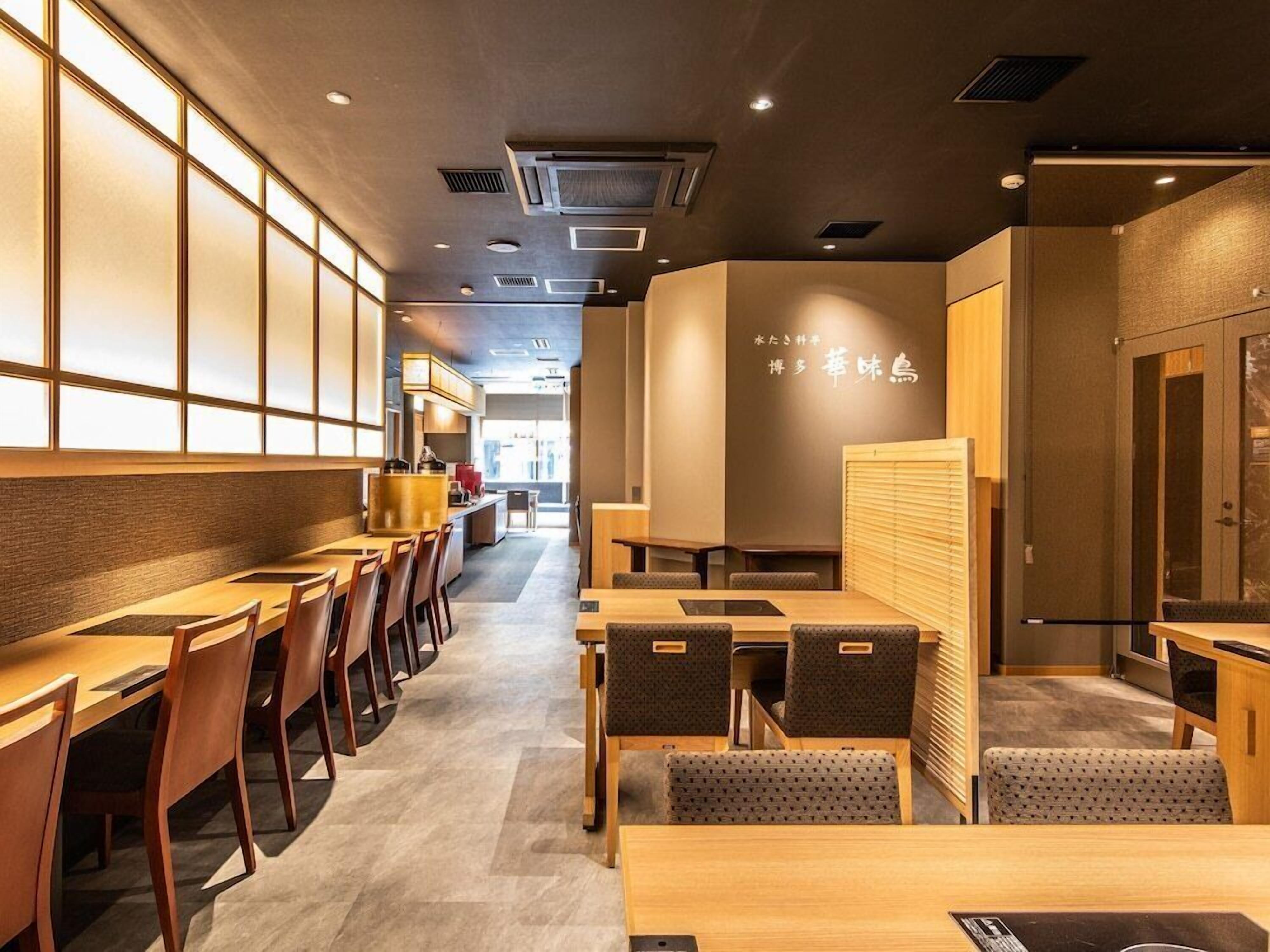 A restaurant interior with wooden tables and chairs, a kitchen area in the background, and a wall with Japanese text.