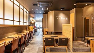 A restaurant interior with wooden tables and chairs, a kitchen area in the background, and a wall with Japanese text.