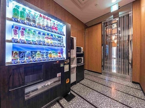 Máquina expendedora con diferentes bebidas y aperitivos, un microondas y una pared de madera en el vestíbulo de un hotel.