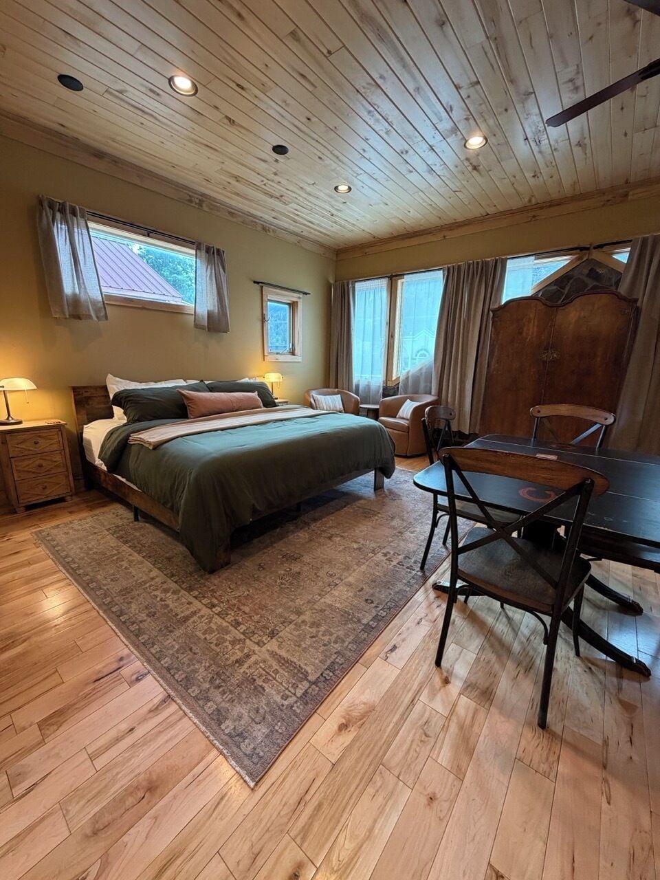 Une pièce avec un plafond en bois, un lit recouvert d’une couverture verte, un bureau en bois avec une chaise et une fenêtre avec des rideaux.