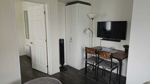 A room with a white door, a black standing heater, a desk with a chair, a TV on the wall, and a floor lamp.