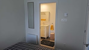 A small, compact room with a bed, a bathroom with a sink and mirror, and a door leading to another room.