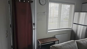 A room with a door, a window with blinds, a clock, a radiator, and a small table.