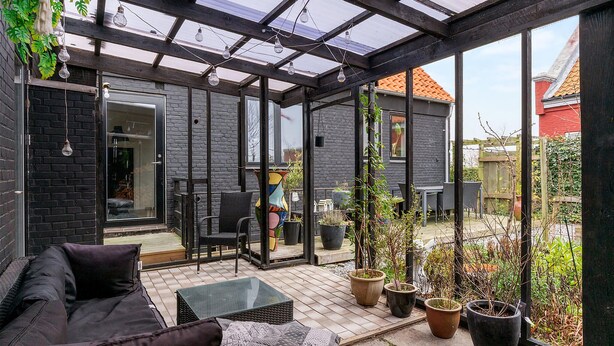 Un patio cubierto con un ventanal que da a una habitación, un sofá, una mesa y plantas en macetas.