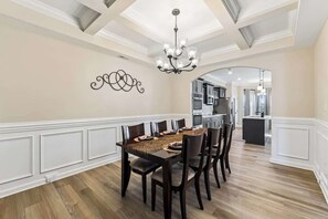 Un coin repas avec une table en bois, des chaises, un lustre et une cuisine en arrière-plan.