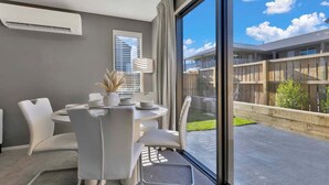 A modern room with a dining table, chairs, a window with a view of a building and greenery, and an air conditioning unit.