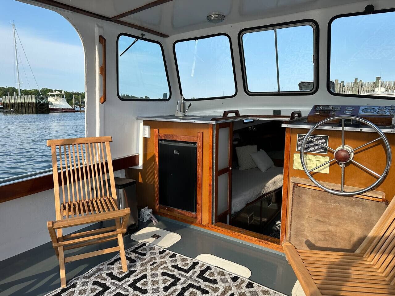 A boat's interior with a wooden chair, a steering wheel, and a small enclosed area with a bed.