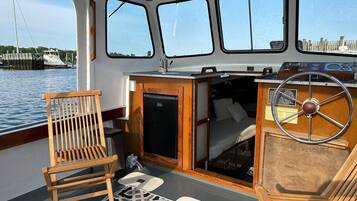 A boat's interior with a wooden chair, a steering wheel, and a small enclosed area with a bed.