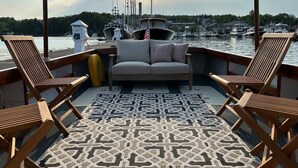 A deck with a patterned rug, wooden lounge chairs, and a sofa facing a marina with boats and a forested hill in the background.