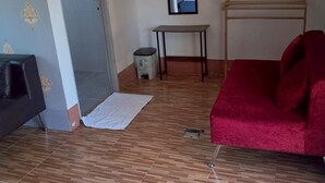A room with a wooden floor, a red sofa, a small table, a mirror, and a door leading to another room. - Phum Riang House (Chaiya)