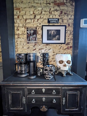 Una estación de café con una máquina Keurig, tazas y objetos con motivos de calaveras frente a una pared de ladrillo con dos cuadros.