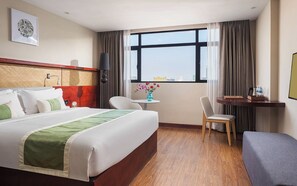 Une chambre d’hôtel avec un grand lit, un bureau, une chaise, une petite table avec un vase, et une vue sur la ville par la fenêtre.