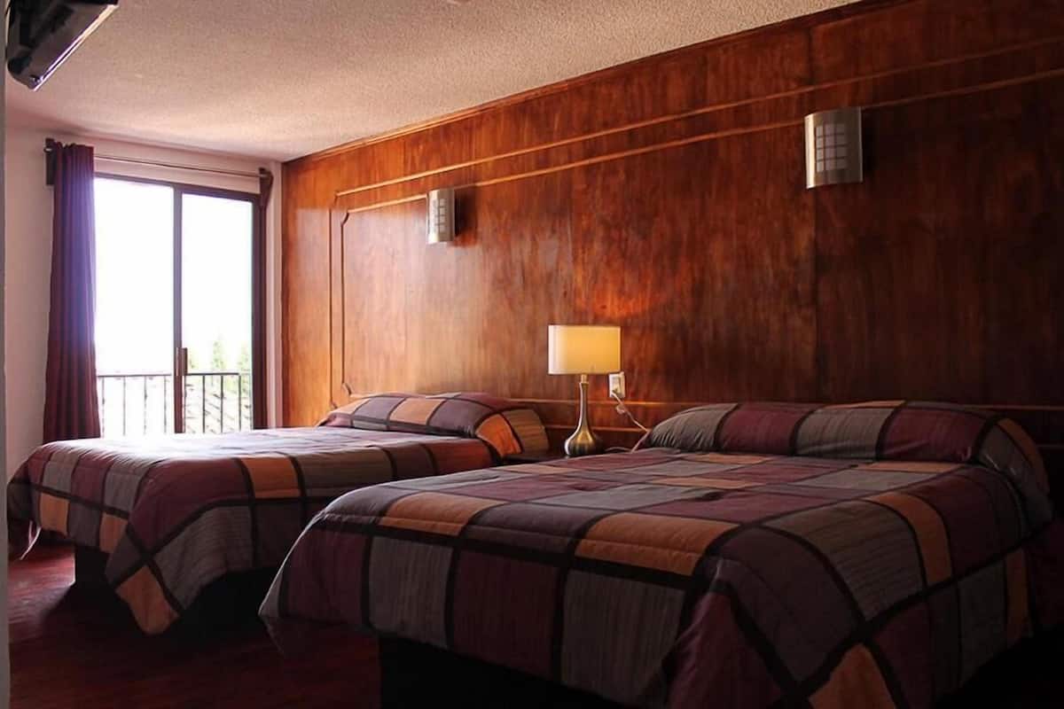 Habitación de hotel con dos camas, una pared de madera, una ventana con cortinas y una mesita de noche con lámpara.