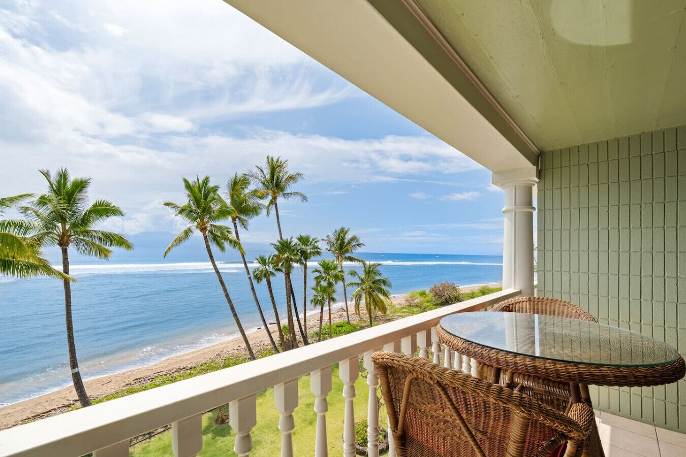 Un balcon doté d’une table en verre et de chaises en osier, offrant une vue sur une plage bordée de palmiers et sur l’océan.
