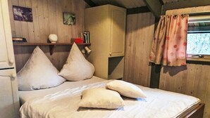 Een slaapkamer met een houten bed, kussens, een nachtkastje, een plank met boeken en een raam met gordijnen.