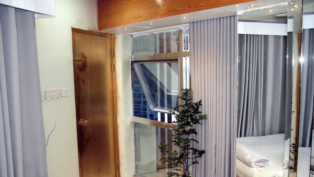 Een kamer met een bed, een kamerplant, een raam met gordijnen, een houten deur en uitzicht op een gebouw.