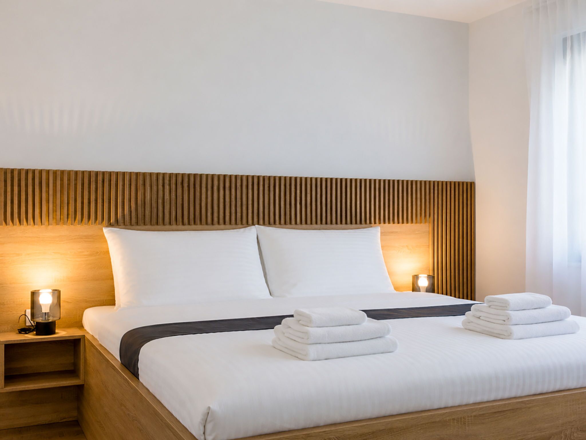 A hotel room with a bed, two bedside lamps, and a stack of towels.