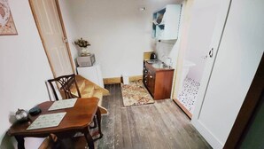 Una habitación estrecha con una mesa de comedor y sillas de madera, una pequeña cocina con un lavamanos y armarios, y un armario blanco.