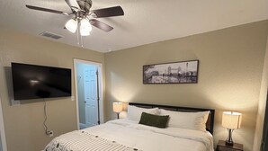 Une chambre à coucher comprenant un lit, un ventilateur de plafond, une télévision, une porte, une table de chevet avec une lampe et un tableau accroché au mur.