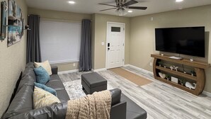 Un salon comprenant un canapé, un pouf, un meuble pour la télévision, un ventilateur de plafond et une porte.