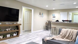 Un salon équipé d’un téléviseur, d’un meuble de rangement en bois, d’un canapé gris et d’un coin cuisine avec un comptoir et des tabourets.