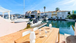 Un port de plaisance avec des bateaux, une terrasse en bois avec des tables et des chaises, et des bâtiments en arrière-plan.