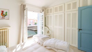 Une chambre à coucher avec un lit, un radiateur blanc, un balcon avec une chaise et une porte dotée de volets à persiennes.