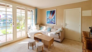 A living room with a sofa, coffee table, and ottomans. There is a painting on the wall and a door to the right. - PORT GRIMAUD: Superb 3-room apartment - A/C, terrace and garden (PORT GRIMAUD)