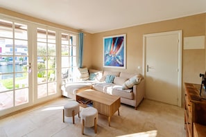 Un salon avec un canapé, une table basse et des poufs. Il y a un tableau au mur et une porte sur la droite.