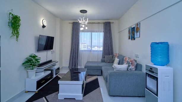 A modern living room with a flat-screen TV, a sofa, a coffee table, a water dispenser, and a ceiling light fixture. - Adorable 1-bedroom bed & breakfast in lively Nairobi (Nairobi)