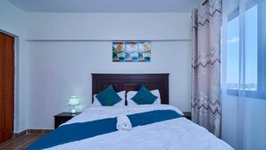 A neatly made bed with a blue and white bedspread, two decorative pillows, a nightstand with a lamp, and a painting above the bed. - Adorable 1-bedroom bed & breakfast in lively Nairobi (Nairobi)