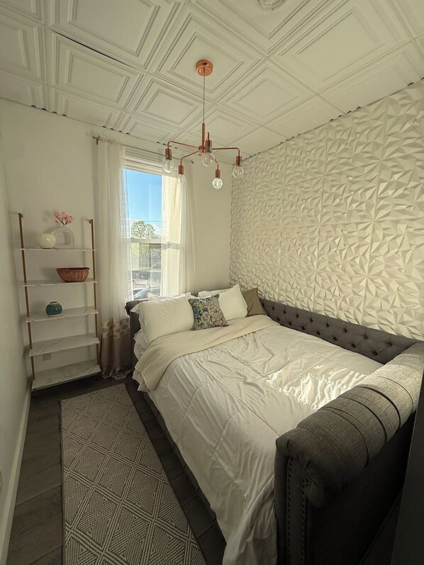 A bedroom with a bed, a shelf with decorative items, a patterned rug, and a textured wall.