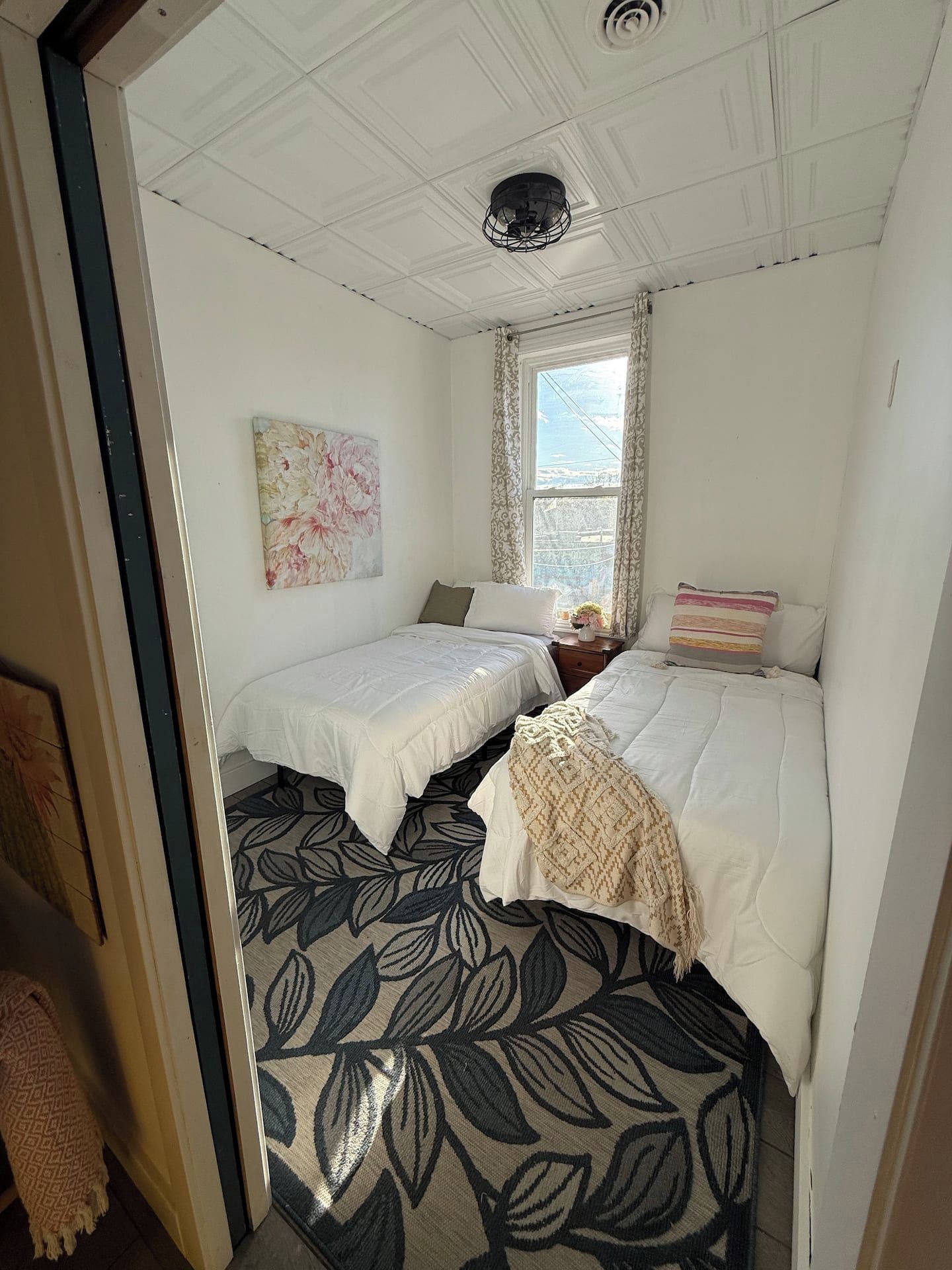 A room with two beds, a window with curtains, a ceiling fan, and a wall-mounted artwork.