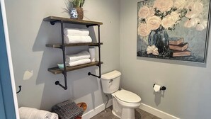 Un baño con un banco de madera, un inodoro, un estante con toallas y un cuadro en la pared.